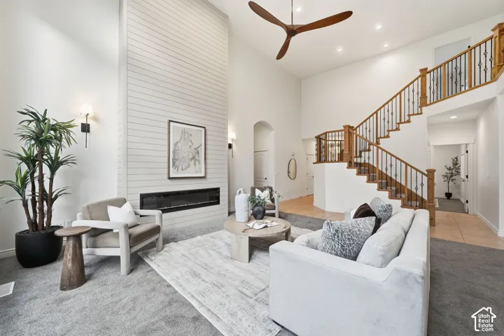 Double-height ceiling, floor-to-ceiling horizontal-plank fireplace surround with built-in linear fireplace, staircase with wood handrail and iron balusters, and multi-blade ceiling fan