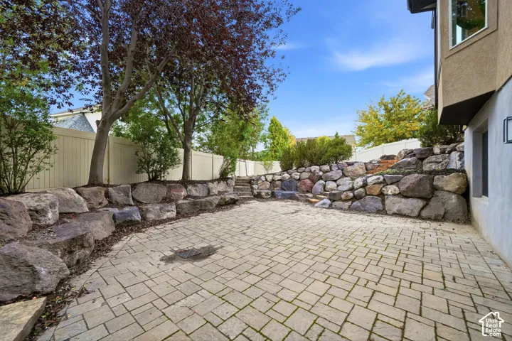 Paver patio featuring multi-level natural stone retaining walls, mature trees, and perimeter vinyl fencing