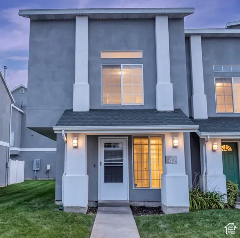 Multi-story exterior featuring a smooth stucco finish, decorative structural columns, dark shingle roofing, and a front entryway with a concrete walkway