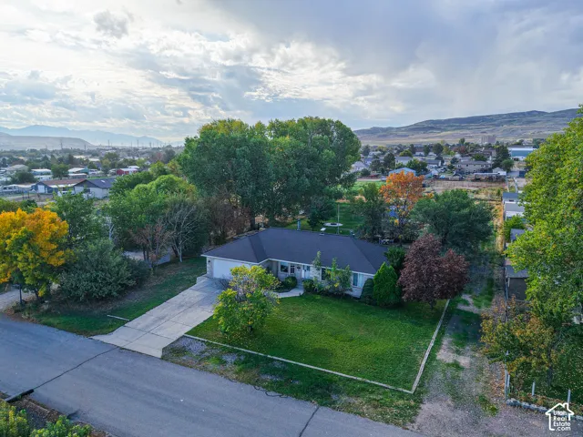 Single-story residence featuring a shingled roof, attached garage, paved driveway, expansive lawn, and mature trees against a mountain backdrop