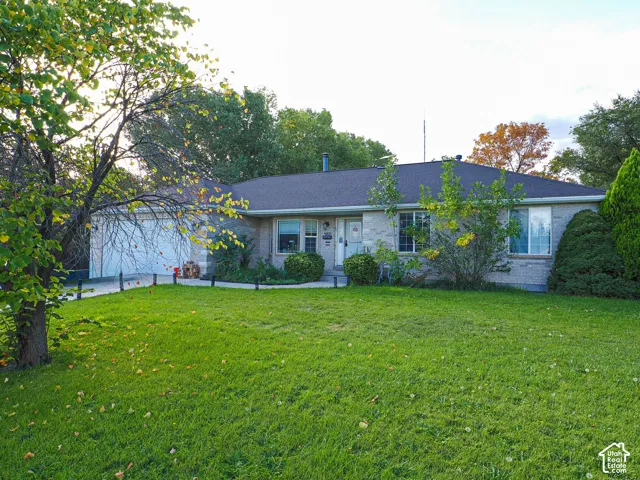 Single-story exterior featuring light-toned masonry, an attached two-car garage, an asphalt shingle roof, and a mature tree-lined front lawn
