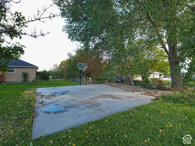 Backyard featuring a concrete sports court with a pole-mounted basketball hoop, grassy lawn areas, and mature trees