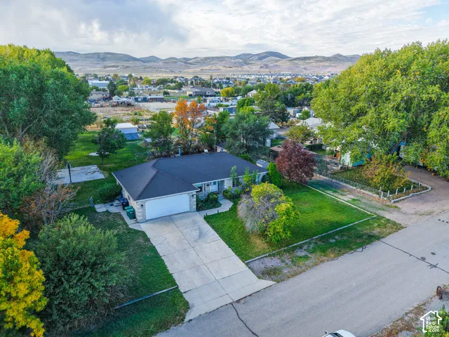 Single-level residence featuring a hipped roof, attached two-car garage, concrete driveway, and expansive lawn with mature trees, set against a mountainous horizon