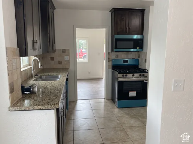Kitchen featuring dark wood cabinetry, natural stone-look countertops, a stone tile backsplash, a stainless steel dual-basin sink with a gooseneck faucet, and ceramic tile flooring