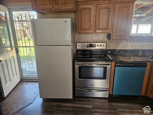 Kitchen featuring wood cabinetry, stainless steel appliance suite, dark stone-finish countertops, tiled backsplash, and wood-finish flooring