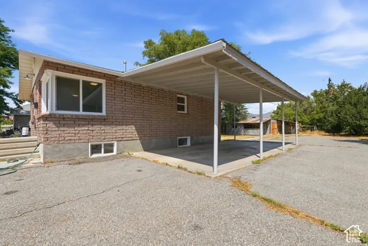 Brick exterior siding, concrete carport with metal support columns, and expansive paved surface