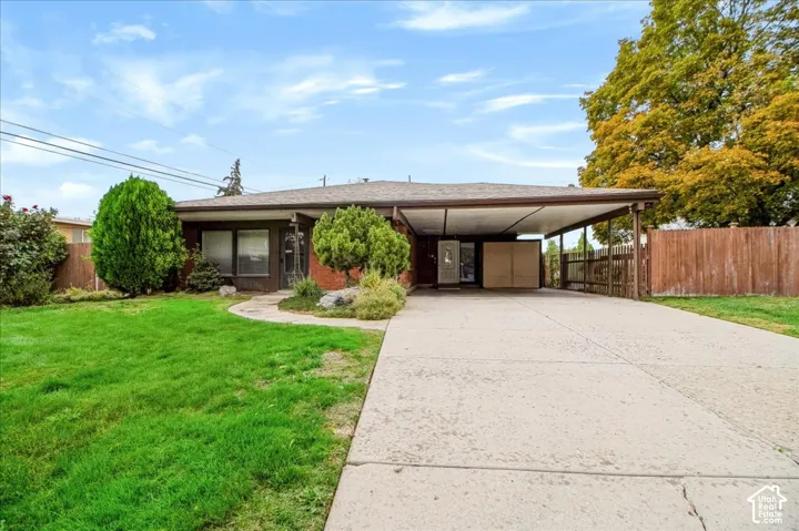 Single-story exterior featuring a brick facade, attached carport, expansive concrete driveway, and established landscaping