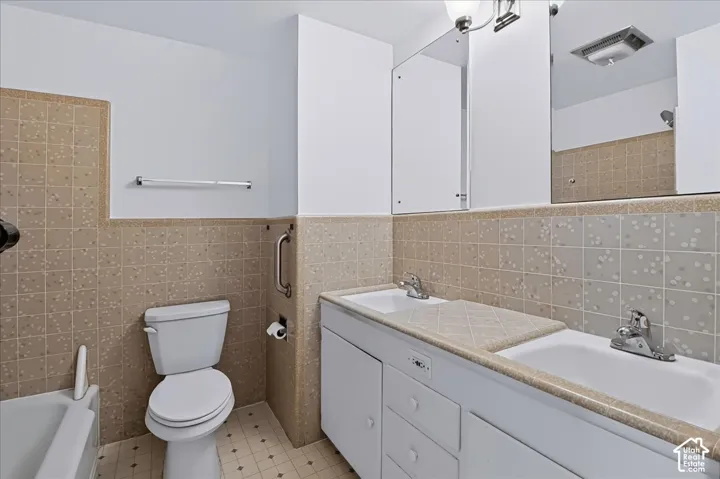 Bathroom featuring a dual-sink vanity with a tiled countertop, white cabinetry, a large wall-mounted mirror, and tile wainscoting