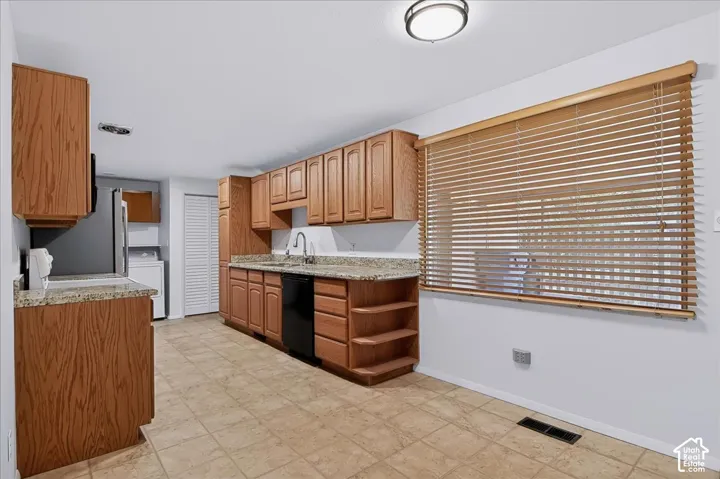 Kitchen featuring natural wood cabinetry, stone-finish countertops, tile flooring, built-in black dishwasher, and large window with horizontal blinds