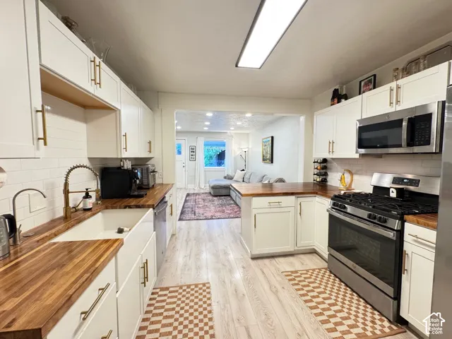Kitchen featuring white shaker-style cabinetry, wood-finish countertops, farmhouse-style apron sink, matte gold-finish hardware, and stainless steel appliances