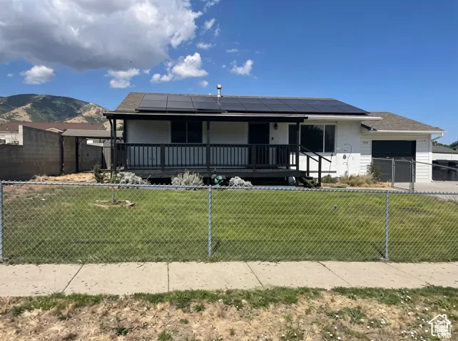 Single-story exterior featuring a covered front deck with railing, roof-mounted solar panels, white siding, and mountain backdrop
