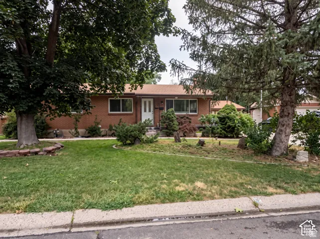 Single-story red brick exterior featuring a shingled roof, mature trees, and landscaped garden beds