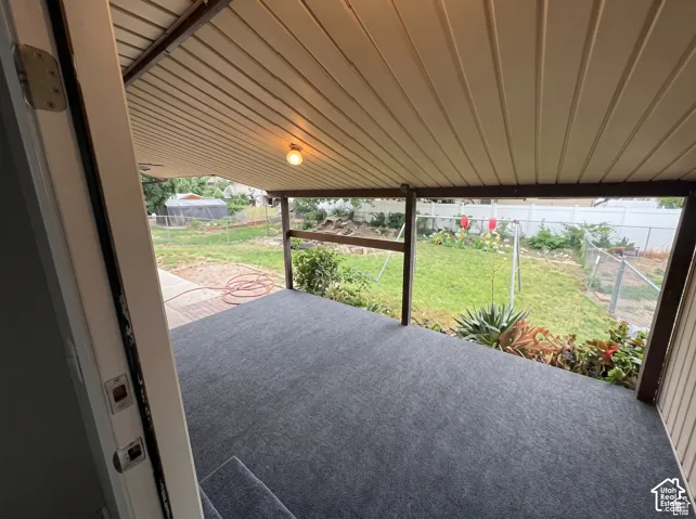 Covered patio featuring carpeted flooring, a ceiling-mounted light fixture, and horizontal beam supports, overlooking a fenced backyard