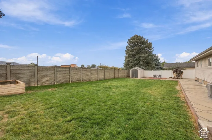 Fenced backyard featuring a manicured lawn, stamped concrete patio, wood-frame garden bed, and a detached storage shed