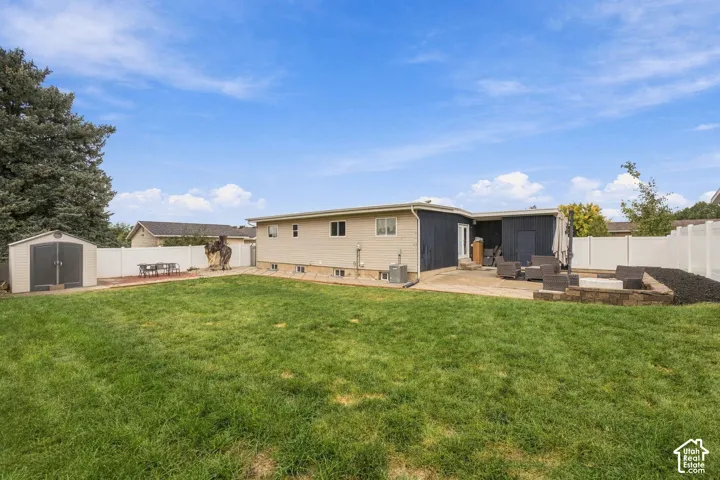Expansive backyard featuring a manicured lawn, concrete patio, stone-block retaining wall, vinyl perimeter fencing, and a detached storage shed