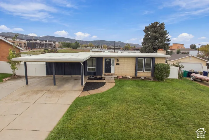 Single-level exterior featuring light-toned masonry, an integrated carport, a detached garage, and a front lawn, set against a mountainous landscape