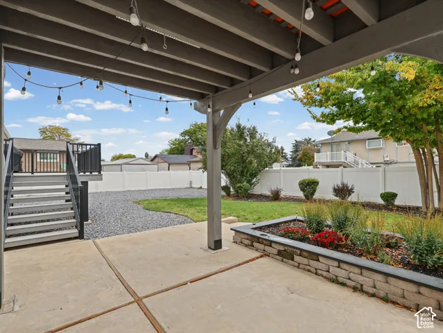 Covered patio featuring exposed wood rafters, concrete flooring, elevated rear deck with black railing, stone-trimmed raised garden bed, and fenced backyard