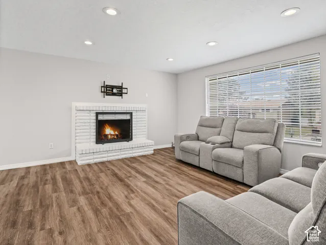 Living area featuring wood-finish flooring, white painted brick fireplace, recessed lighting, and large window with horizontal blinds