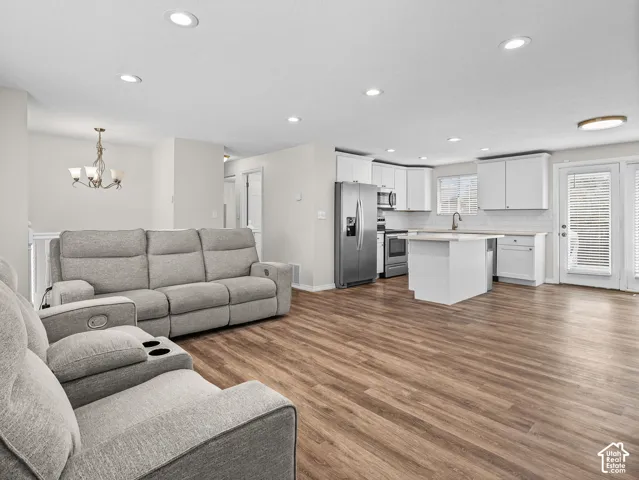 Open-concept living and kitchen space featuring wood-finish flooring throughout, a gray upholstered reclining sectional sofa, stainless steel appliances including a refrigerator and range, white cabinetry with a center island, recessed ceiling lighting, a