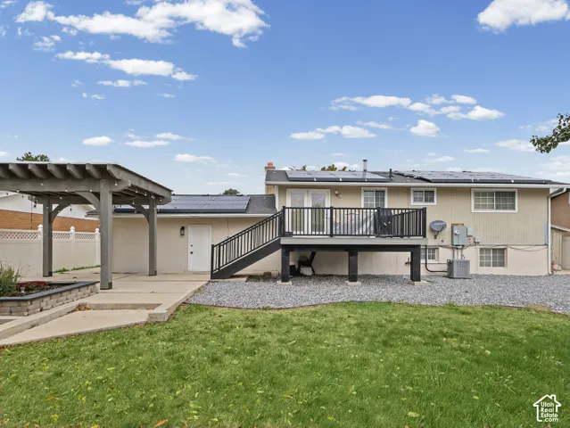 Backyard featuring an elevated deck with metal railing, a detached wooden pergola, a poured concrete patio, solar panel roof installation, and perimeter vinyl fencing