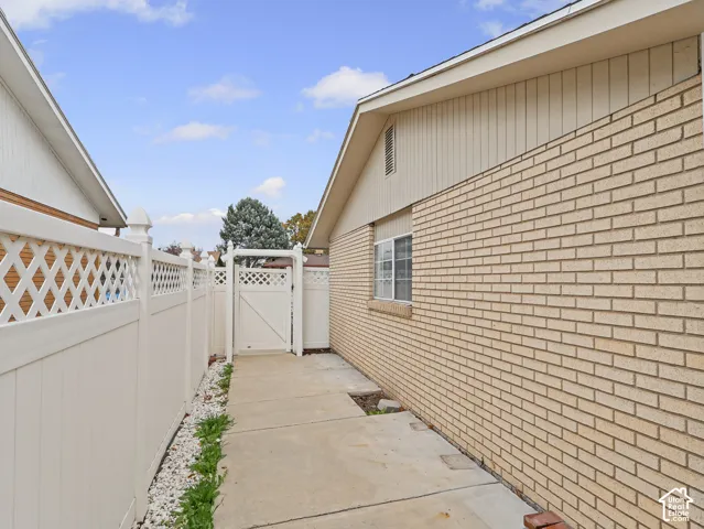 Exterior featuring tan brick siding, vertical paneled siding, a concrete side path, and white vinyl privacy fencing with integrated lattice and a matching gate