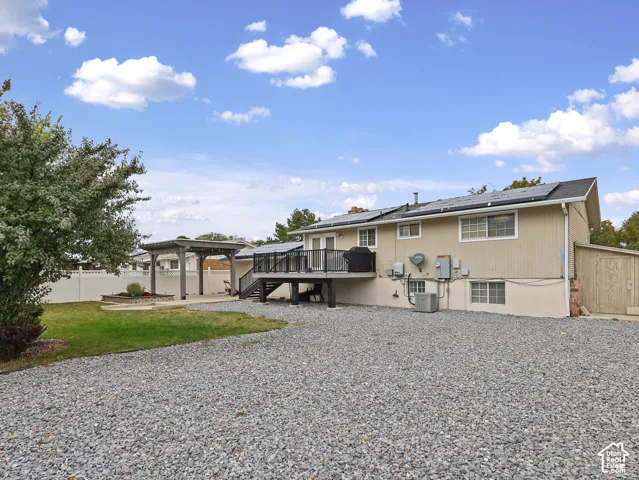 Exterior rear elevation featuring roof-mounted solar panels, an elevated deck with black metal railing, a detached pergola, and gravel ground cover