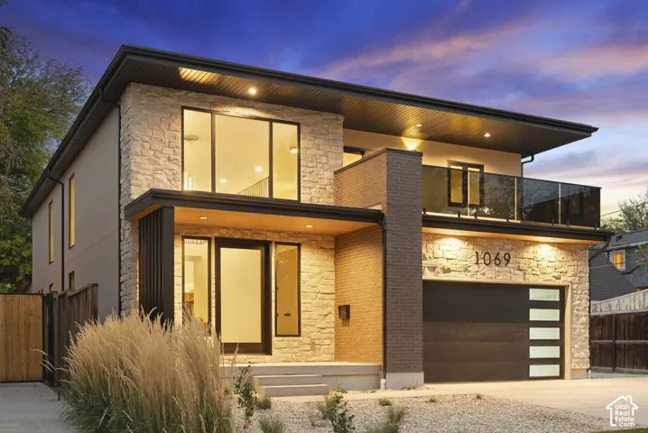 Contemporary two-story exterior featuring multi-textured stone and brick masonry, large-format glass windows, a covered entryway with wood-finish ceiling detailing, and an attached balcony with a glass railing