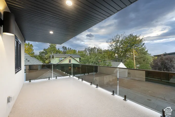 Covered balcony featuring a low-maintenance stone-composite deck surface, glass railing with metal standoffs, recessed ceiling lighting, and wall-mounted exterior light fixtures