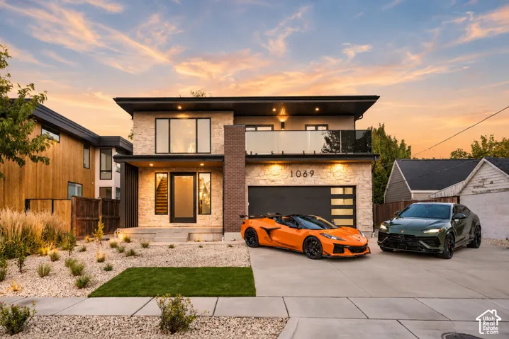 Modern exterior featuring stone and brick masonry, a second-level balcony with glass railing, large-pane windows, and a multi-panel garage door