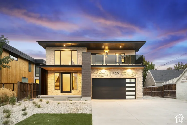 Modern exterior featuring natural stone veneer, dark brick accents, a glass-railing second-story balcony, and a garage door with horizontal glass panels
