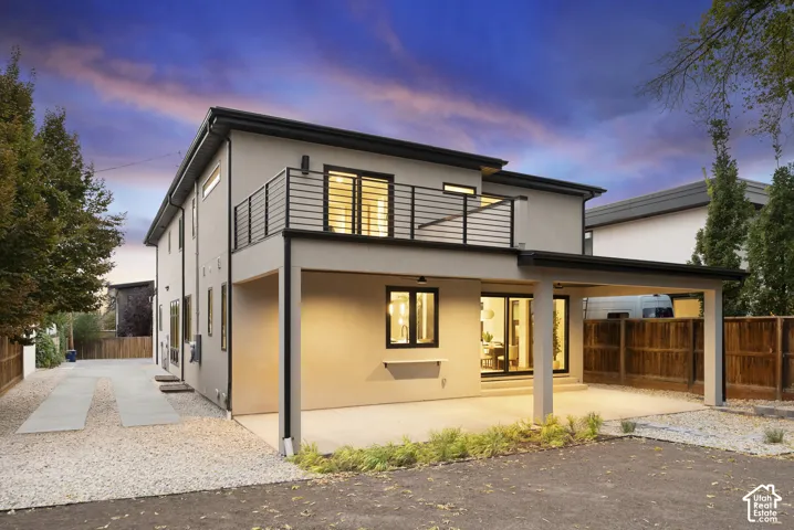 Stucco exterior featuring an upper-level balcony with horizontal metal railings, a covered lower patio, sliding glass doors, and dark-framed windows