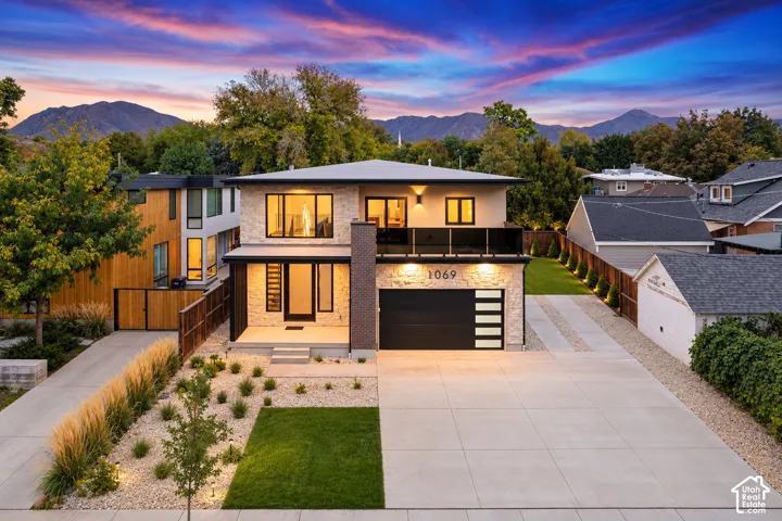 Contemporary two-story exterior featuring stone veneer siding, a flat roofline, second-level balcony with glass railing, and a multi-panel sectional garage door