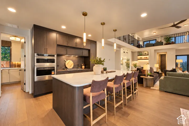 Kitchen featuring flat-panel dark wood cabinetry, expansive stone-surface island, built-in double wall ovens, wood-finish flooring, and minimalist pendant lighting