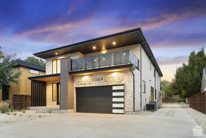 Contemporary exterior featuring a textured stone facade, integrated garage with frosted horizontal panels, upper-level glass balcony, and recessed soffit lighting