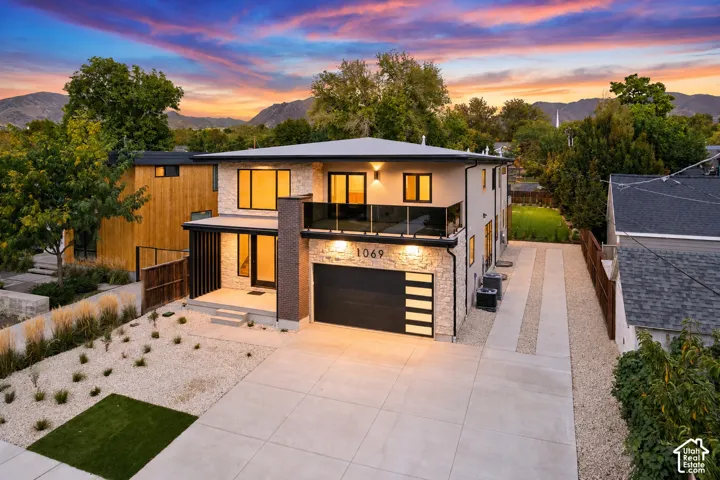 Modern multi-level exterior featuring stacked stone masonry, a glass-railed balcony, an attached two-car garage with horizontal light panels, and a concrete driveway