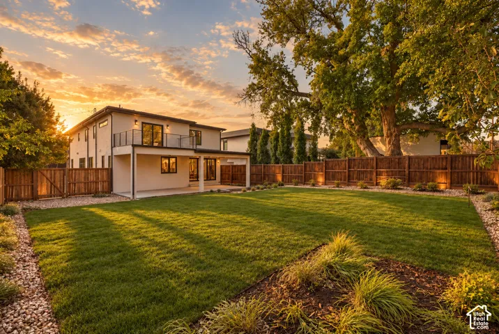 Expansive backyard featuring a manicured lawn, stone-lined landscape beds, a covered patio, and a second-story balcony with metal railings, all enclosed by a wooden privacy fence