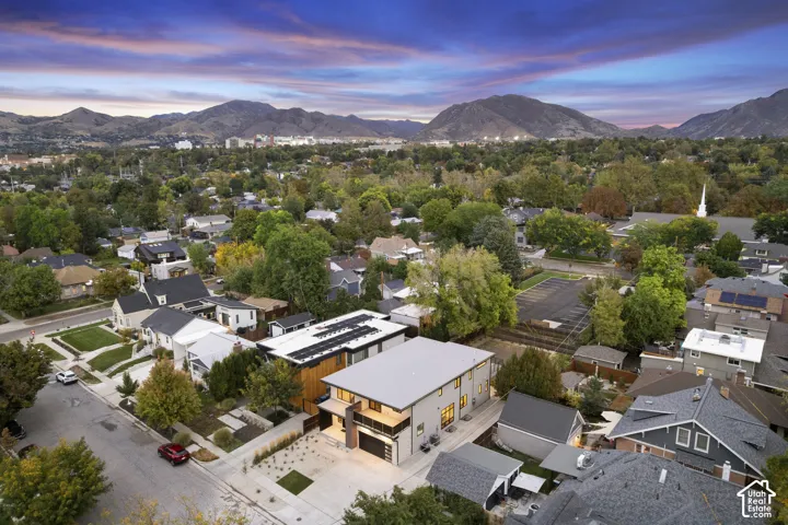 Aerial view of a contemporary residence featuring a flat roof with integrated solar panels, a two-story architectural design, a private balcony with railing, and a concrete driveway, set against a backdrop of mountain scenery