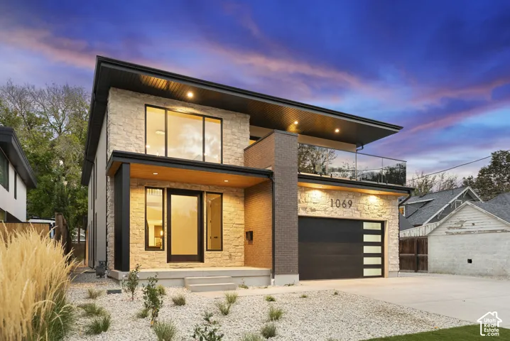 Modern two-story exterior featuring a stacked-stone facade, glass-panel balcony railing, recessed lighting, and a multi-panel garage door
