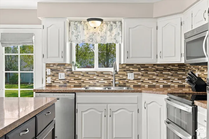Kitchen featuring white cabinetry, stone-finish countertops, decorative multi-toned tile backsplash, stainless steel appliances, and a double-basin sink