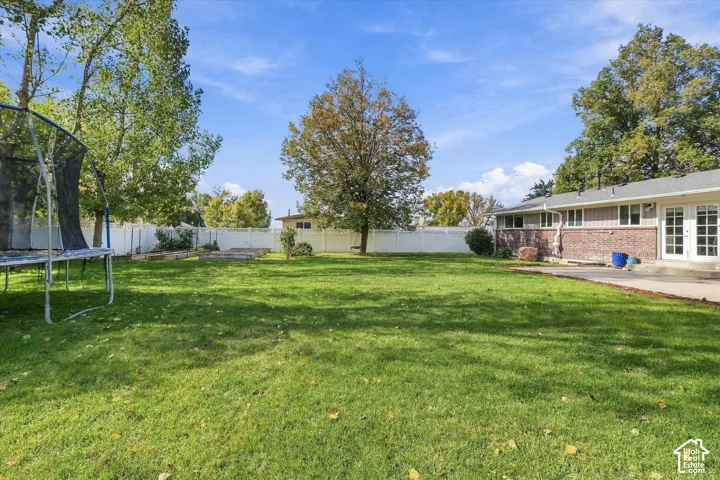 Expansive backyard featuring a large grass lawn, perimeter white vinyl fencing, mature trees, raised garden beds, and a concrete patio