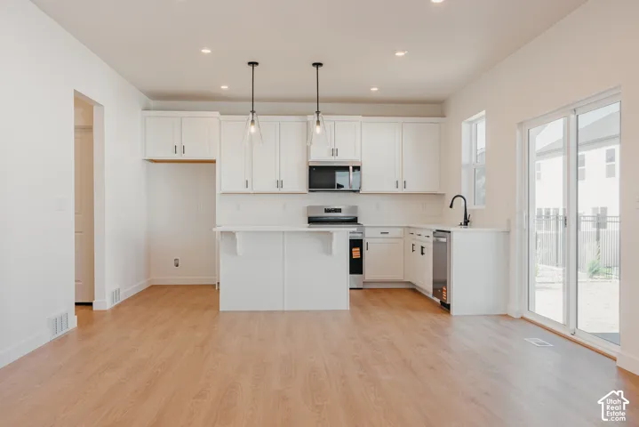 Kitchen featuring white shaker-style cabinetry, wood-finish flooring, a central island with a breakfast bar, stainless steel appliances, and dual pendant lighting