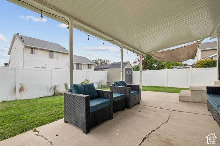 Covered concrete patio with overhead string lighting, enclosed by a white vinyl privacy fence, including a grassy lawn and a detached storage shed
