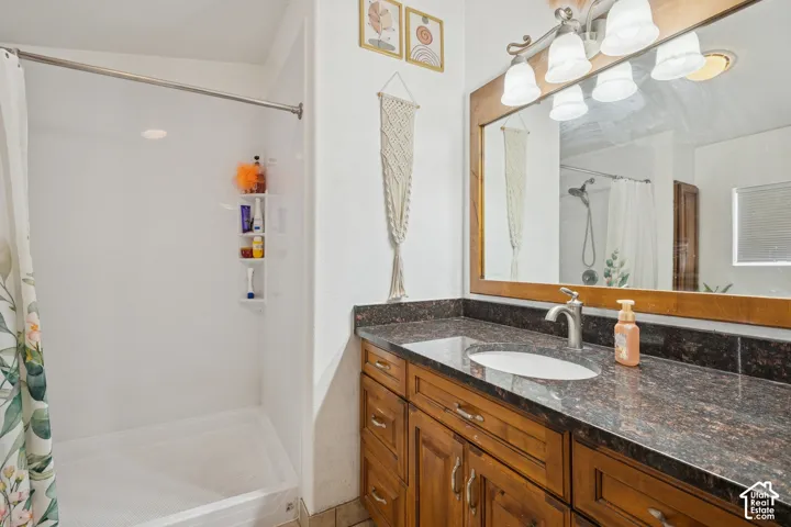Bathroom featuring a vanity with wood-finish cabinetry, a dark stone-finish countertop, an oval under-mount sink, a full-width mirror with a wood frame, and a shower stall with an integrated surround