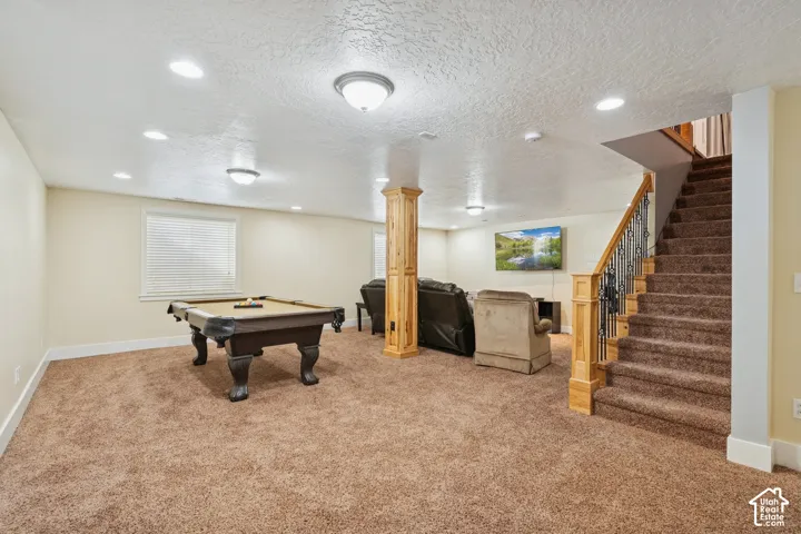 Open-concept space featuring recessed lighting, carpet flooring, a central wood-encased support column, and a staircase with ornate metal balusters and a wood handrail