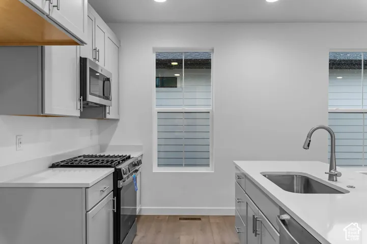 Kitchen featuring white cabinetry, solid-surface countertops, stainless steel appliances including a gas range and built-in microwave, a center island with an undermount sink and pull-down faucet, and wood-finish flooring