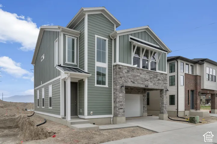 Two-story exterior featuring sage green vertical and horizontal siding, stone veneer accents, an integrated garage, and a metal roof portico