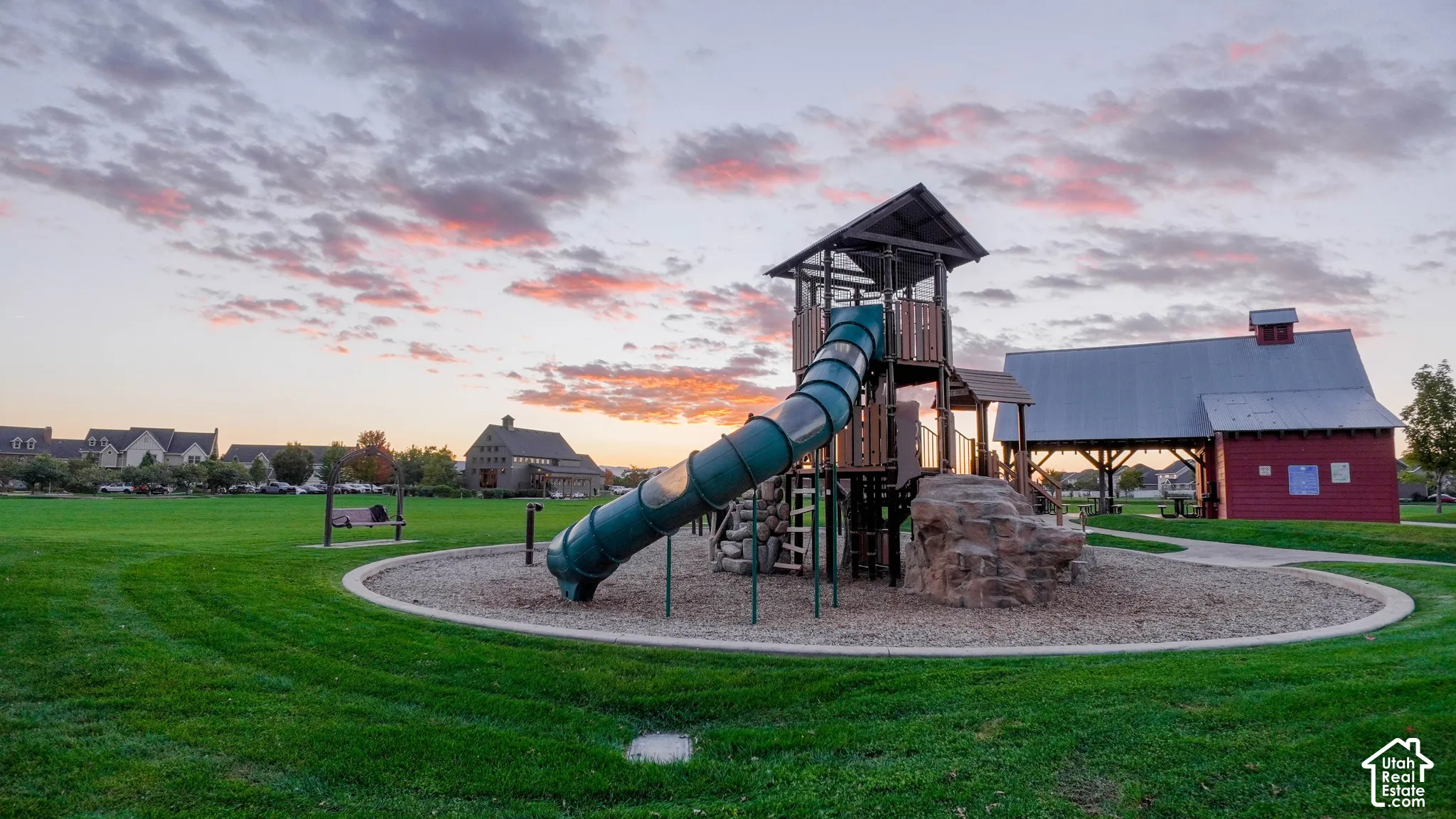 Expansive park grounds featuring a multi-level playground structure with a slide, climbing element, wood-chip base, and a red barn-style pavilion under a mountain backdrop