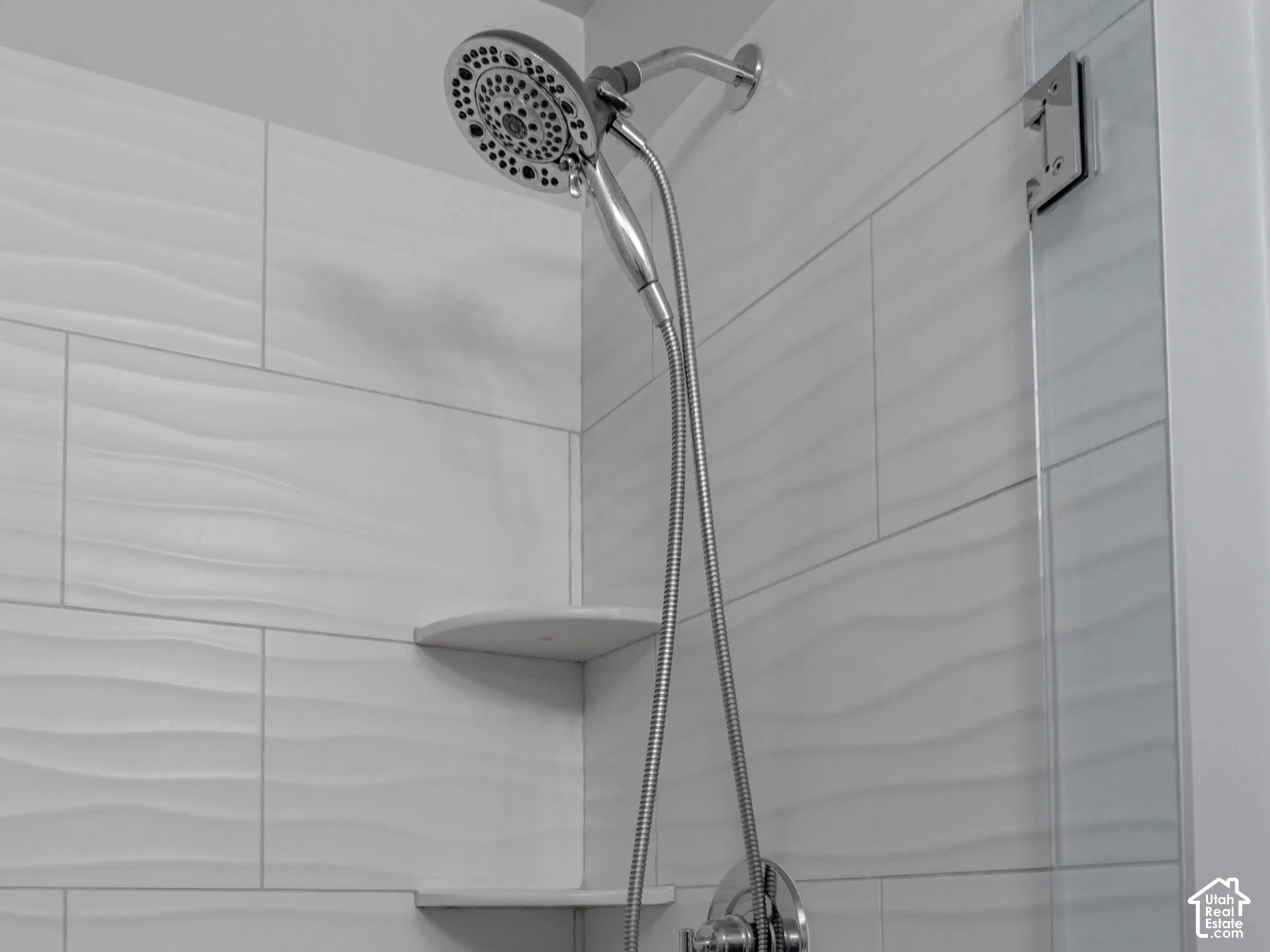 Shower enclosure featuring textured white wall tile, dual integrated corner shelving, a chrome handheld shower head, and a glass partition