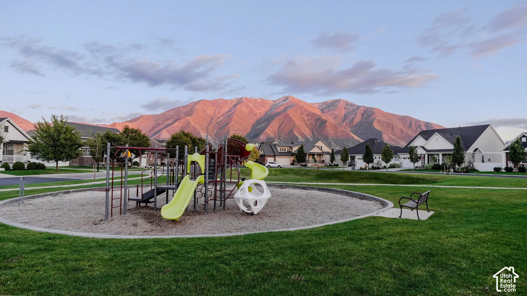 Community park featuring a playground structure with slides, manicured lawn, metal bench, and mountain backdrop