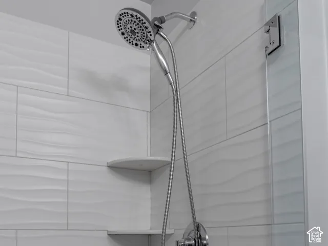 Shower enclosure featuring textured white wall tile, dual integrated corner shelving, a chrome handheld shower head, and a glass partition